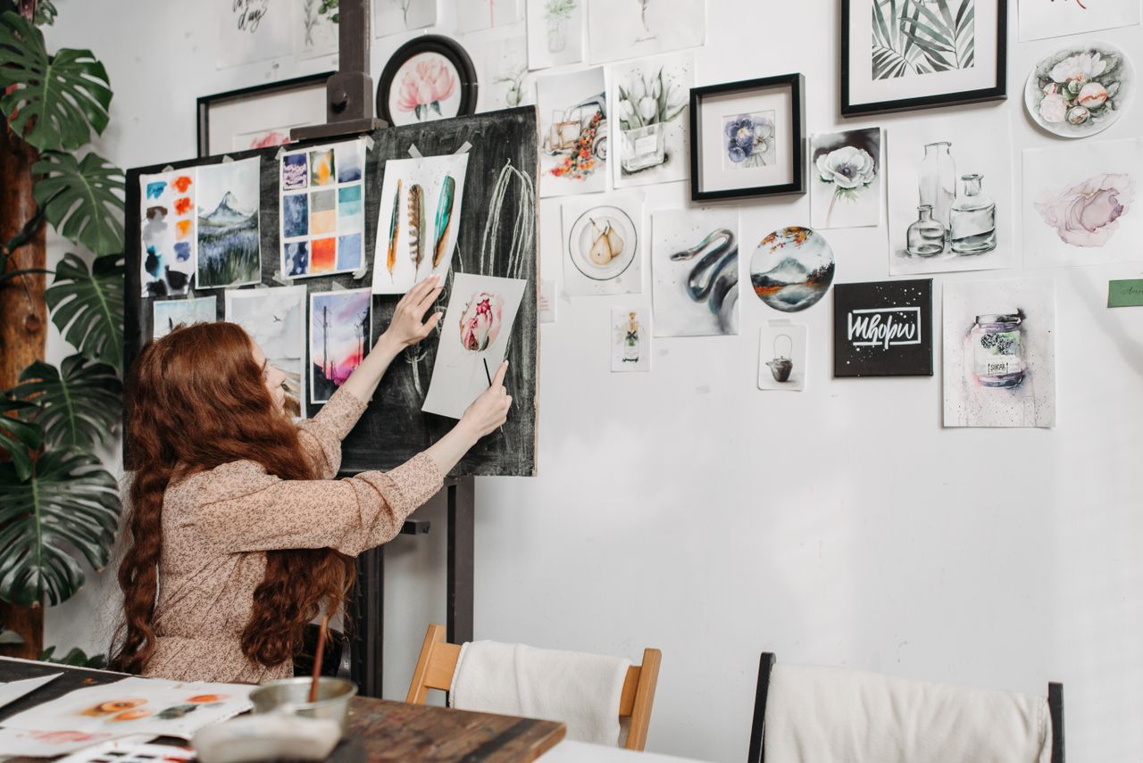 about-us-02 Woman artist organizing colorful paintings on a gallery wall. Creative and inspiring studio environment.