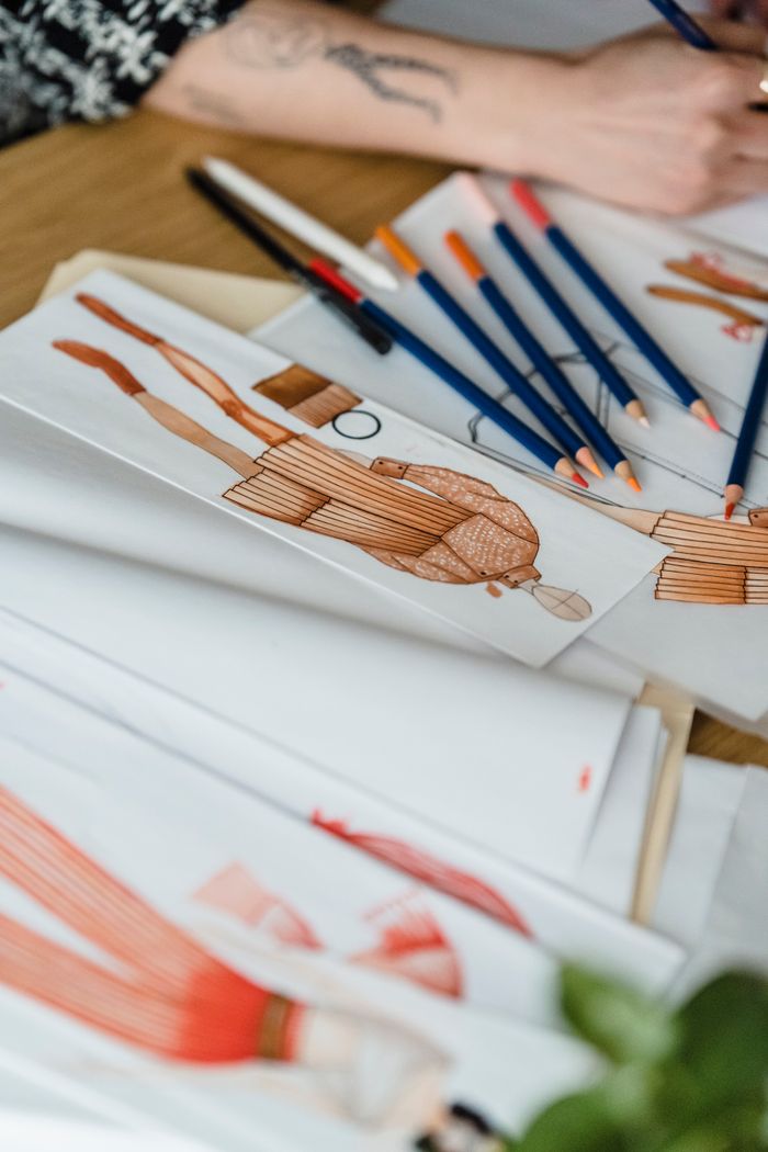 services-04 From above of crop anonymous female designer at desk with pencils drawing against illustrations of human figures in outfit