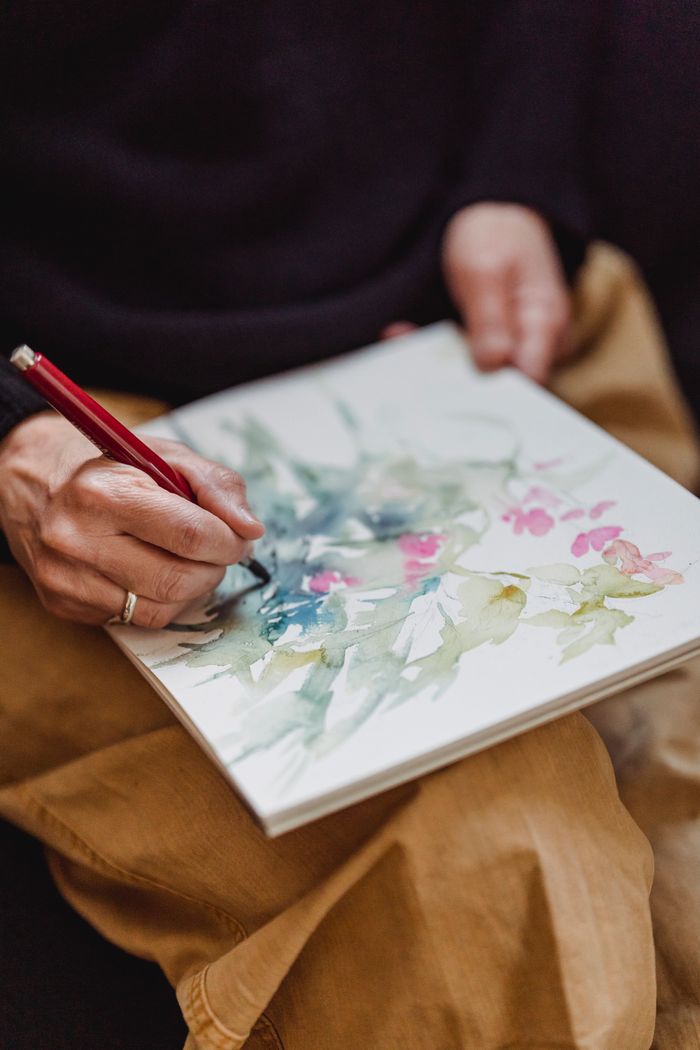 about-us-02 Close-up of hands painting delicate watercolor flowers and foliage in a sketchbook.